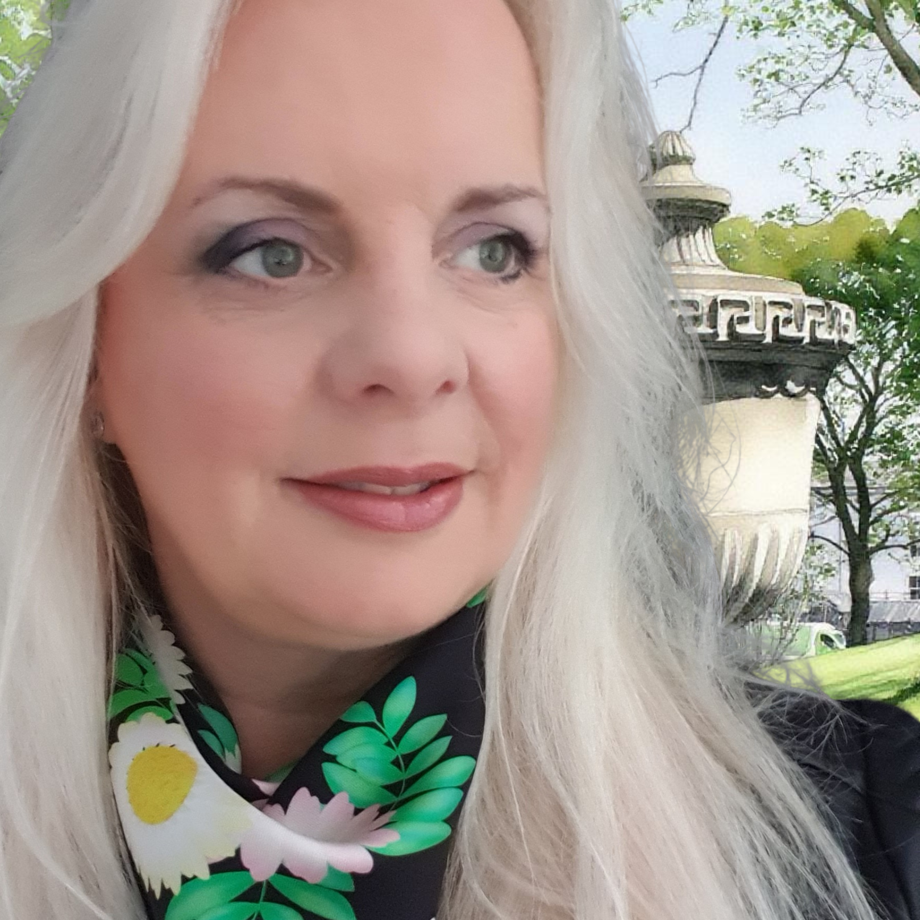 Woman with long white hair wearing a floral scarf, outdoors with trees and a lamp post in the background.