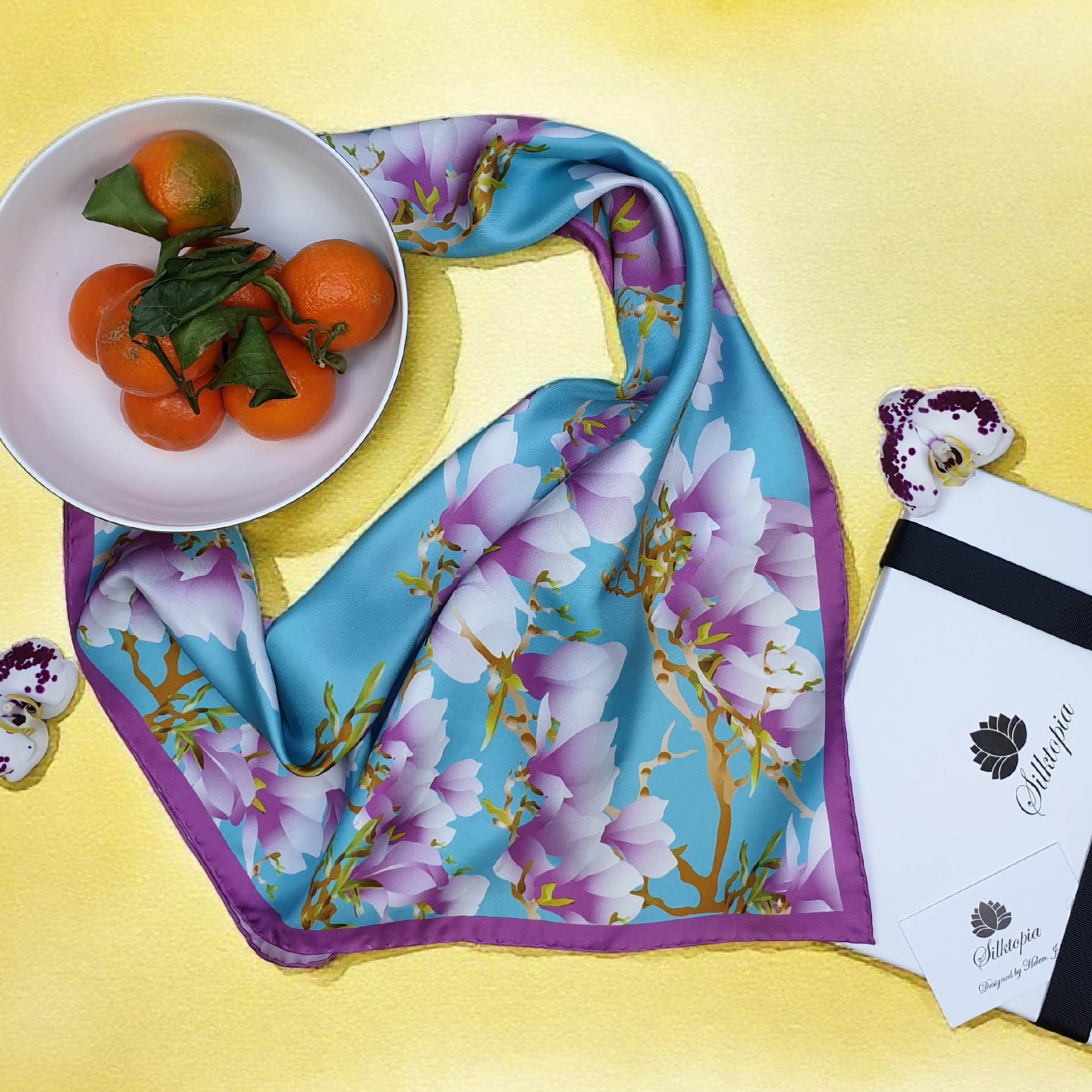 Floral scarf on a yellow surface with a bowl of oranges and a card.